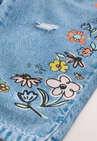 Világoskék farmeranyag, színes virágos hímzéssel rózsaszín, narancs és sárga árnyalatokban. A anyagon kopott részlet is található.