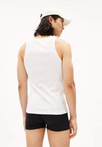 Witte katoenen tanktop met een aansluitend ontwerp, gezien vanaf de achterkant, gecombineerd met zwarte shorts, op een modell met lang donker haar.