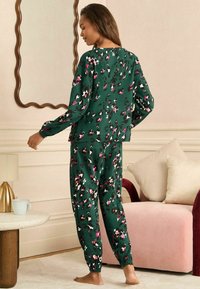 Roheline pyjama komplekt, millel on laia lõikega särk ja kitsenevad püksid, kaunistatud lillemustriga roosa, valge ja musta värviga. Pehme kangastekstsuur.