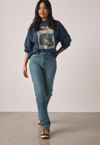 Tumesinine dressipluus lehma graafikaga ja kuldse tekstiga, kombineeritud helesiniste sirge lõikega teksade ning pruunide ja kreemjate tossudega.