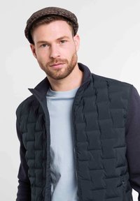 Navy quilted vest with zip closure and high collar, layered over a light blue t-shirt. Model wearing a brown flat cap.