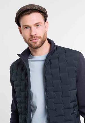 Navy quilted vest with zip closure and high collar, layered over a light blue t-shirt. Model wearing a brown flat cap.
