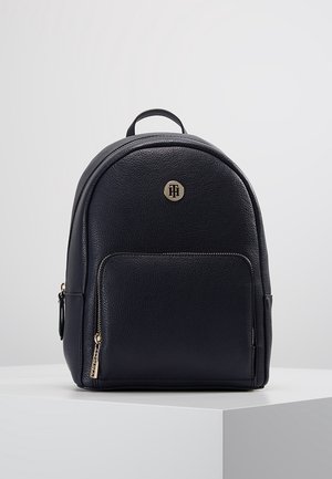 Sac à dos en cuir noir avec poche avant zippée, tirette de fermeture éclair dorée, petite poignée supérieure et emblème rond en métal avec logo sur fond lisse.