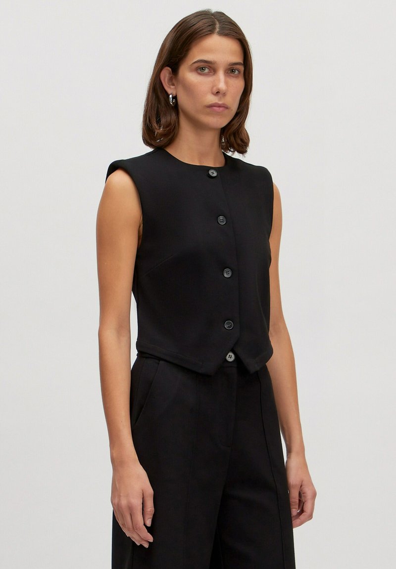 Zwarte mouwloze top met een ronde hals en een sluiting met vijf knopen aan de voorkant. De stof heeft een gladde textuur, een getailleerde fit en een cropped ontwerp.
