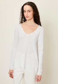 Ženska s kodrastimi lasmi, oblečena v belo majico z dolgimi rokavi in V-izrezom ter svetle hlače, stoji pred enobarvnim svetlim ozadjem.