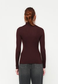 Frau mit rotem Haar im Dutt, die ein enges, gerippte, langärmliges, kastanienbraunes Oberteil und lockere, dunkelschwarze Jeans trägt, steht vor einer schlichten weißen Wand.
