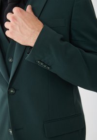 Ciemnozielona marynarka o gładkiej fakturze, z wypustkami na klapach, trzema guzikami na mankietach i kieszenią na piersi.