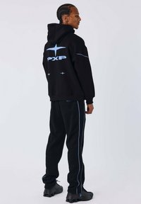 Hombre con el cabello trenzado que lleva una sudadera negra con el logo azul "PXP" y pantalones negros a juego con rayas laterales azules, de pie contra un fondo claro.