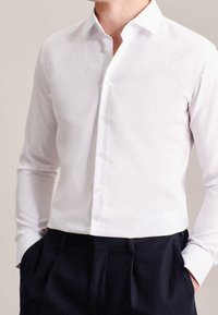Camicia bianca a maniche lunghe con colletto a punta e chiusura frontale con bottoni, caratterizzata da una texture liscia e un design aderente, abbinata a pantaloni scuri.