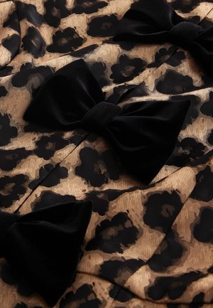 Čierna a zlatá látka s leopardím vzorom, zdobená veľkými sametovými mašľami. Materiál má textúrovaný, tkaný vzor, ktorý vytvára vizuálnu hĺbku a kontrast.