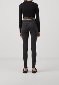 Vrouw in een zwart langmouw cropped top en donkergrijze skinny jeans, staande op blote voeten op een effen lichte achtergrond.