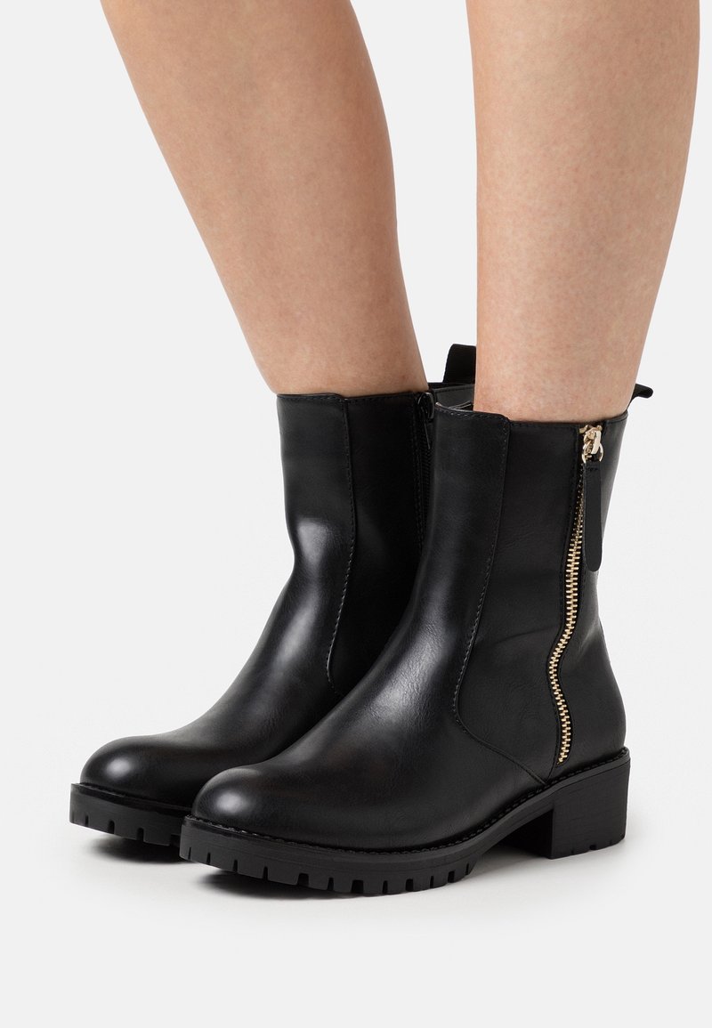Bottines en cuir noir avec semelle épaisse, petit talon et fermeture éclair latérale dorée portées sur jambes nues sur fond blanc.