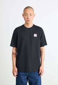T-shirt en coton noir à manches courtes, col rond, avec un petit patch logo rectangulaire sur la poitrine gauche. Porté avec un jean en denim bleu.