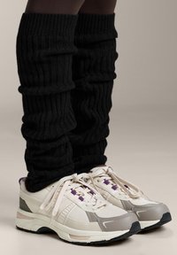 Černé žebrované návleky na nohy zakrývají dolní část nohou, doplněné lehkými teniskami s šedou a bílou síťovinou a fialovými detaily.