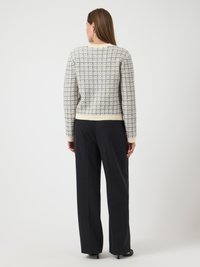 Γυναίκα που στέκεται στρα�μμένη προς τα πίσω, φορώντας ένα κρεμ και μπλε καρό πουλόβερ και μαύρους παντελόνες με φαρδιά πόδια και μαύρες γόβες.