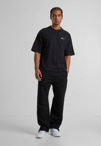 Camiseta negra de gran tamaño con un pequeño logo en el pecho, combinada con pantalones de chándal negros y zapatillas blancas, que muestran un ajuste casual y relajado.