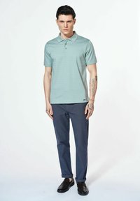 Miętowa koszula polo z delikatnym wzorem w paski, w połączeniu z granatowymi spodniami. Model ma na sobie czarne buty, prezentując tatuaże na ramionach.