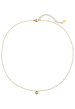 Vrouw in een wit colbert draagt een bijpassende gouden hartvormige hangerketting en een delicaat armbandje, hand rustend op de borst.