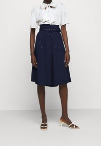 Gonna blu navy con chiusura frontale a bottoni, tasche laterali e cintura larga; abbinata a una camicia bianca con colletto a fiocco. Sandali con tacco.