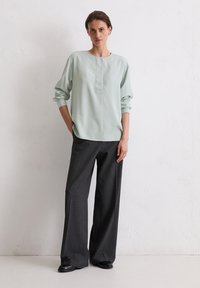 Hellgrüne, langärmlige Bluse mit rundem Ausschnitt und einer Knopfleiste vorne; kombiniert mit dunkelgrauen, weit geschnittenen Hosen und schwarzer Schuhen.