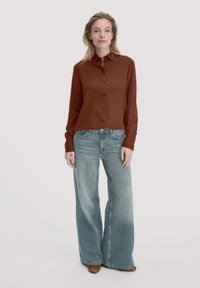 Camicia a maniche lunghe color ruggine con bottoni, abbinata a jeans a gamba larga azzurri e scarpe marroni, su uno sfondo neutro.