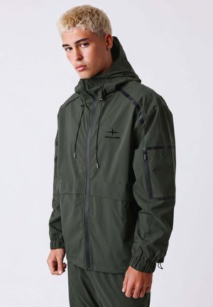 Jeune homme aux cheveux blonds courts portant une veste à capuche vert foncé avec une poche zippée sur la manche et des cordons noirs, debout devant un fond uni.