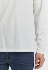 Langærmet hvid skjorte lavet af blødt stof, med lige snit, der har sideslidser i kanten og et lille mærke på sidebanen.