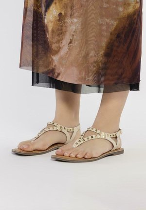 Sandalias beige con acentos de tachuelas, con un diseño de tiras en T y suela plana. Las tiras son suaves, con una estética minimalista.