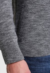 Šedý pletený svetr s žebrovanými manžetami a spodním lemem, s texturovaným vzorem. Viditelná ruka spočívající na tmavé džínové látce.