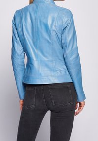 Kvinde iført en tætsiddende lyseblå læderjakke og sorte denimjeans, set fra ryggen mod en ensfarvet hvid baggrund.