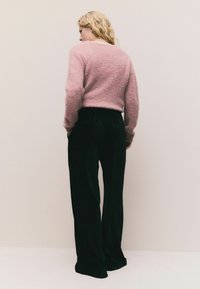 Lyserød, struktureret sweater med lange ærmer, parret med sorte, løstsiddende bukser med elastisk talje. Ensfarvet baggrund.