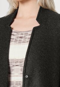 Mörkgrå filtjacka med rund hals och tryckknappsstängning, lager på lager över en ljus mönstrad stickad topp med horisontella r änder.