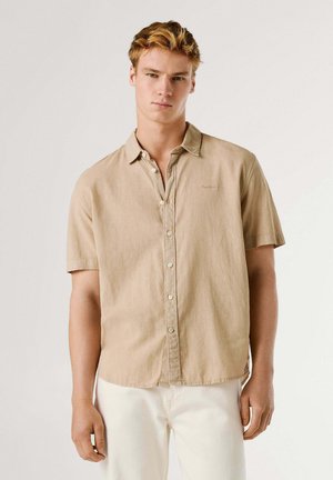 Jonge man met kort blond haar, gekleed in een beige overhemd met korte mouwen en witte broek, staand tegen een effen achtergrond.