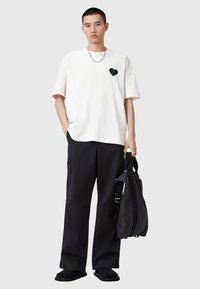 Camiseta blanca con un parche de corazón negro, combinada con pantalones cargo negros y zapatillas negras sin cordones, sosteniendo una bolsa negra con correas para el hombro.