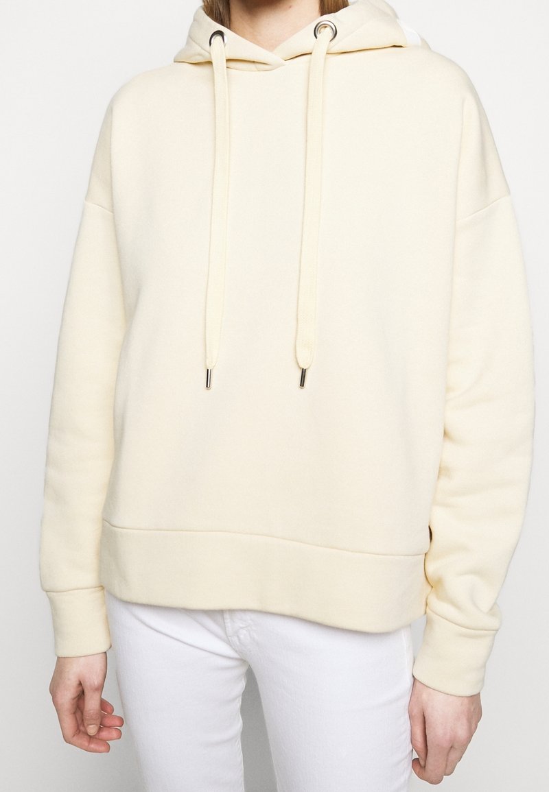 Personne portant un sweat à capuche crème avec cordons et un pantalon blanc, mettant l'accent sur le haut du corps et les mains sur les côtés.