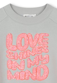 Sweatshirt gris clair avec un col rond arborant un grand texte rose en sequins qui dit "L'AMOUR BRILLE DANS MON ESPRIT." Texture douce.