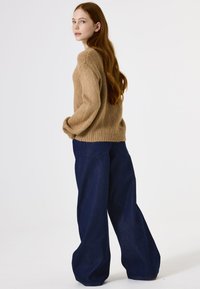 Brauner Mohair-Mix-Pullover mit strukturierter Oberfläche und gerippten Bündchen, kombiniert mit weit geschnittenen, dunkelblauen Jeanshosen mit Gesäßtaschen.