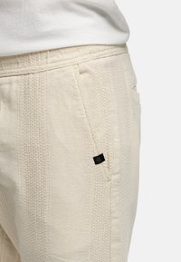 Cremfarbene Hosen aus strukturiertem Gewebe, mit vertikalen Mustern, seitlichen Taschen und einem kleinen schwarzen Logo-Label am Bund.