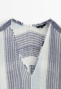 Chemise en tissu léger à rayures verticales bleu marine et blanches avec des bords effilochés autour du col et de l'encolure.