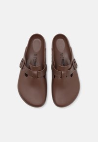 Scarpe marroni, slip-on, con una superficie liscia in plastica, dotate di una piccola fibbia e fori di ventilazione sui lati.