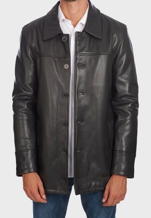 Mand iført en sort læderjakke med knapper over en hvid skjorte og blå jeans, stående mod en ensfarvet baggrund.