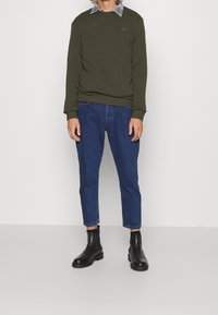 Sudadera verde oliva con puños acanalados, combinada con jeans azul ajustados y botas negras de tobillo. Detalle discreto del logo en el pecho.