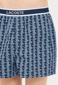 Șorturi de baie bleumarin, cu un model geometric de culoare albastră deschisă. Talie elastică cu logo-ul "LACOSTE" și închidere cu buton în față.
