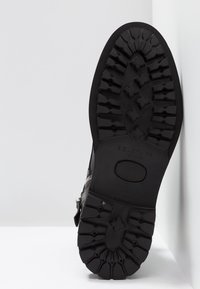 Bota de cuero negro que muestra una suela de goma rugosa y texturizada con grandes tacos de tracción y un detalle de cremallera lateral. Nombre de la marca grabado visible.