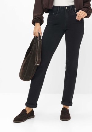 Person iført sort slim-fit jeans med op-rullede manchetter, mørkebrune ruskind loafers og holder en matchende mørkebrun ruskindstaske.