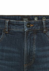 Donkerblauwe denim jeans met oranje stiksels, voorzien van een knopen sluiting en twee voorzakken. Het label toont "Camel Active."