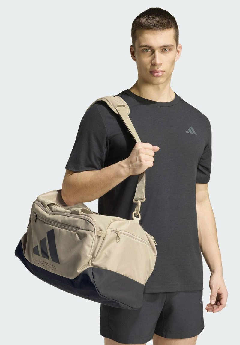 Jeune homme portant une tenue de sport noire, portant un sac de sport Adidas beige et noir sur l'épaule sur un fond uni.