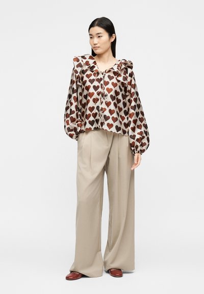 Γυναίκα που φοράει μια φαρδιά μπλούζα με σχέδιο καρδιές και με βολάν στον γιακά, μπεζ παντελόνια με φαρδιά μπατζάκια και καφέ παπούτσια, στέκεται μπροστά σε απλό φόντο.