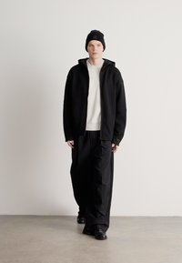 Μαύρο τζάκετ με φερμουάρ πάνω από λευκό ριμπ πλεκτό πουλόβερ, συνδυασμένο με φαρδιά μαύρη παντελόνια και μαύρο σκουφί. Ομαλές υφές, άνετη εφαρμογή.