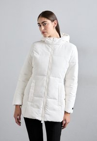 Žena nosi bijelu puffer jaknu s kapuljačom, koja ima prednje džepove i potpuni zatvarač, stojeći naslonjena na jednostavnu svijetlosivu pozadinu.